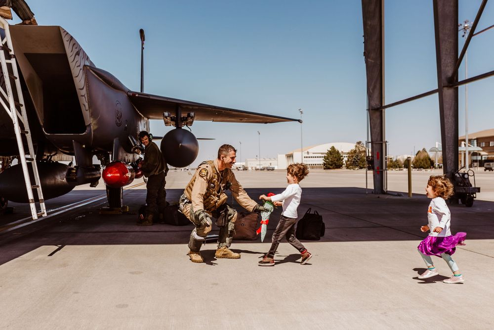 Military service member kneels by aircraft on tarmac as two young children play nearby at airbase.