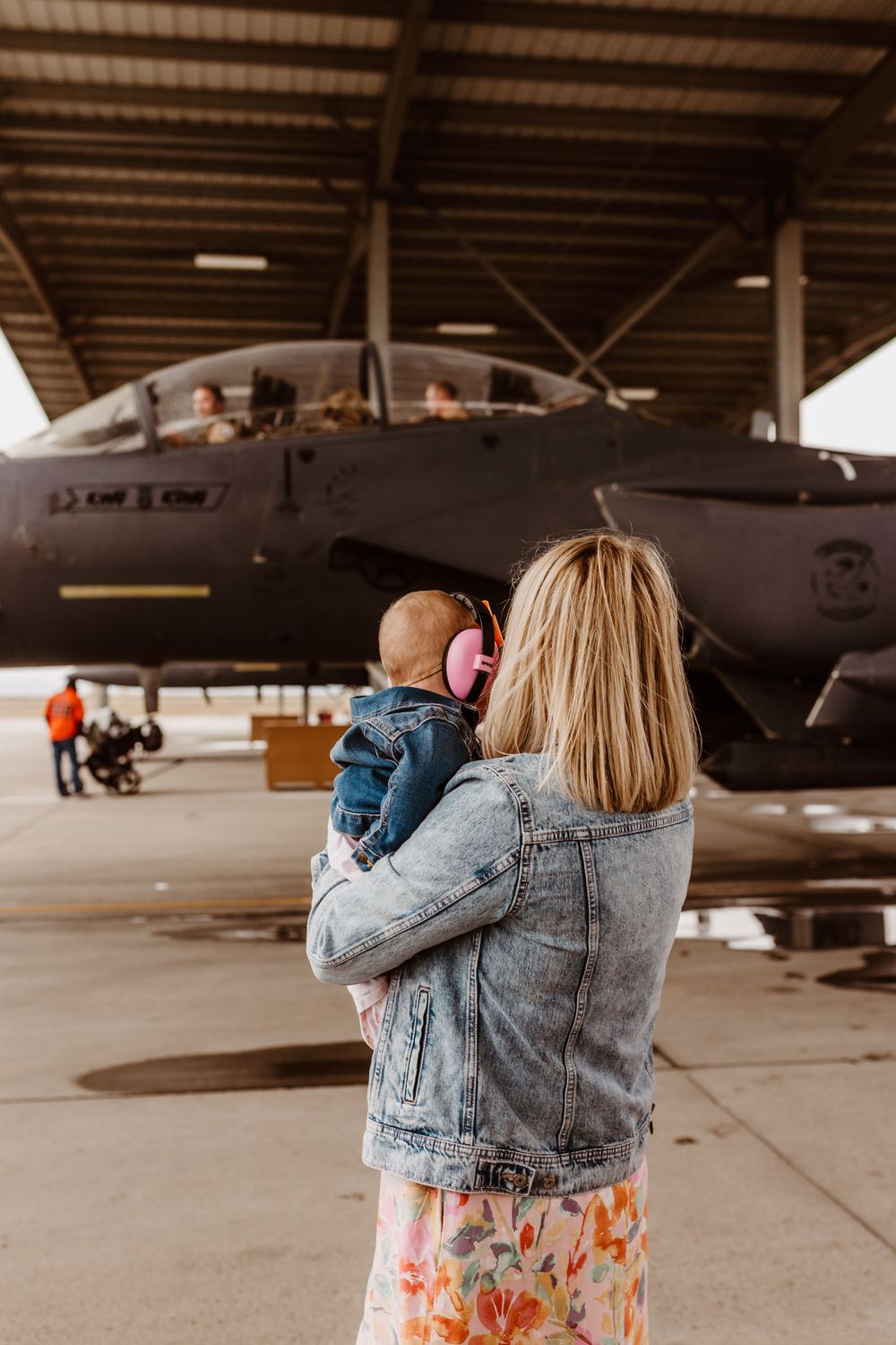 A parent wearing a denim jacket holds a small child while looking at a military aircraft in an indoor hangar. Military homecoming.