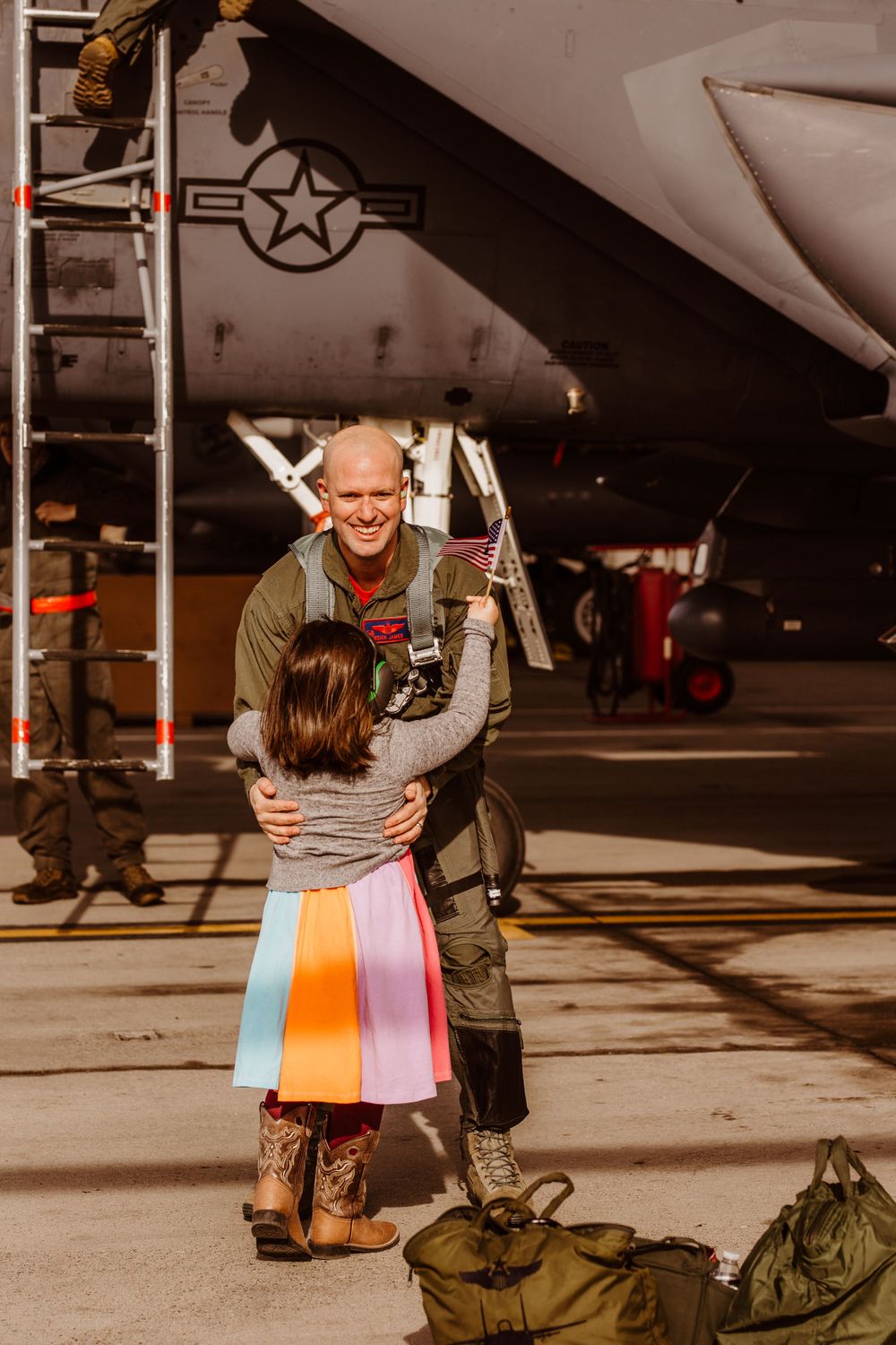 Military personnel in flight suit embraces loved one on airfield next to military aircraft with duffel bags on ground.