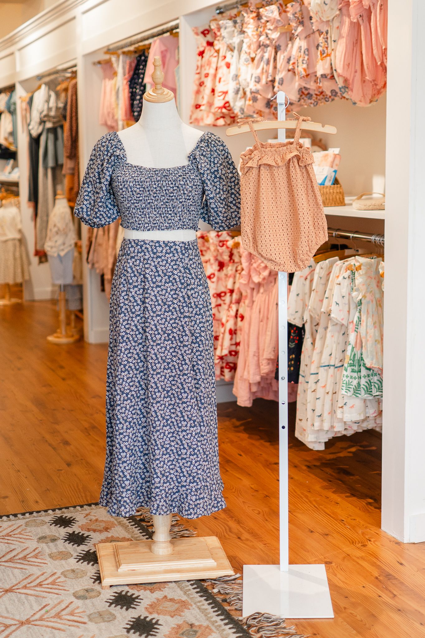 A blue floral maxi dress with puff sleeves on display in a boutique with pink and peach clothing racks in background.