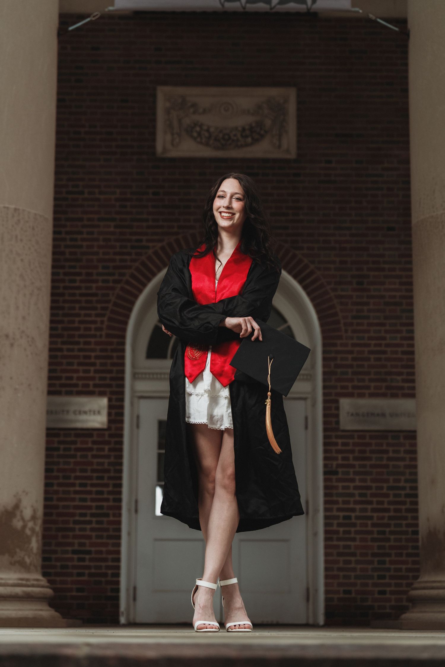 Smiling graduate in black gown and red sash holds cap outside brick building with white columns in Cincinnati