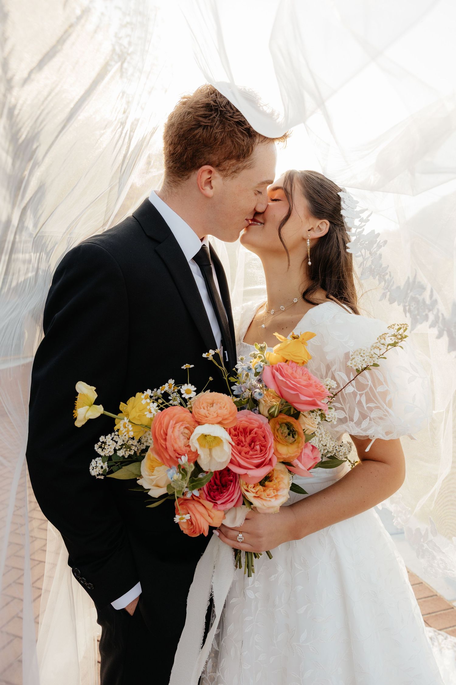 A bride and groom kiss under a flowing veil, bride holding a colorful bouquet of pink and yellow flowers.