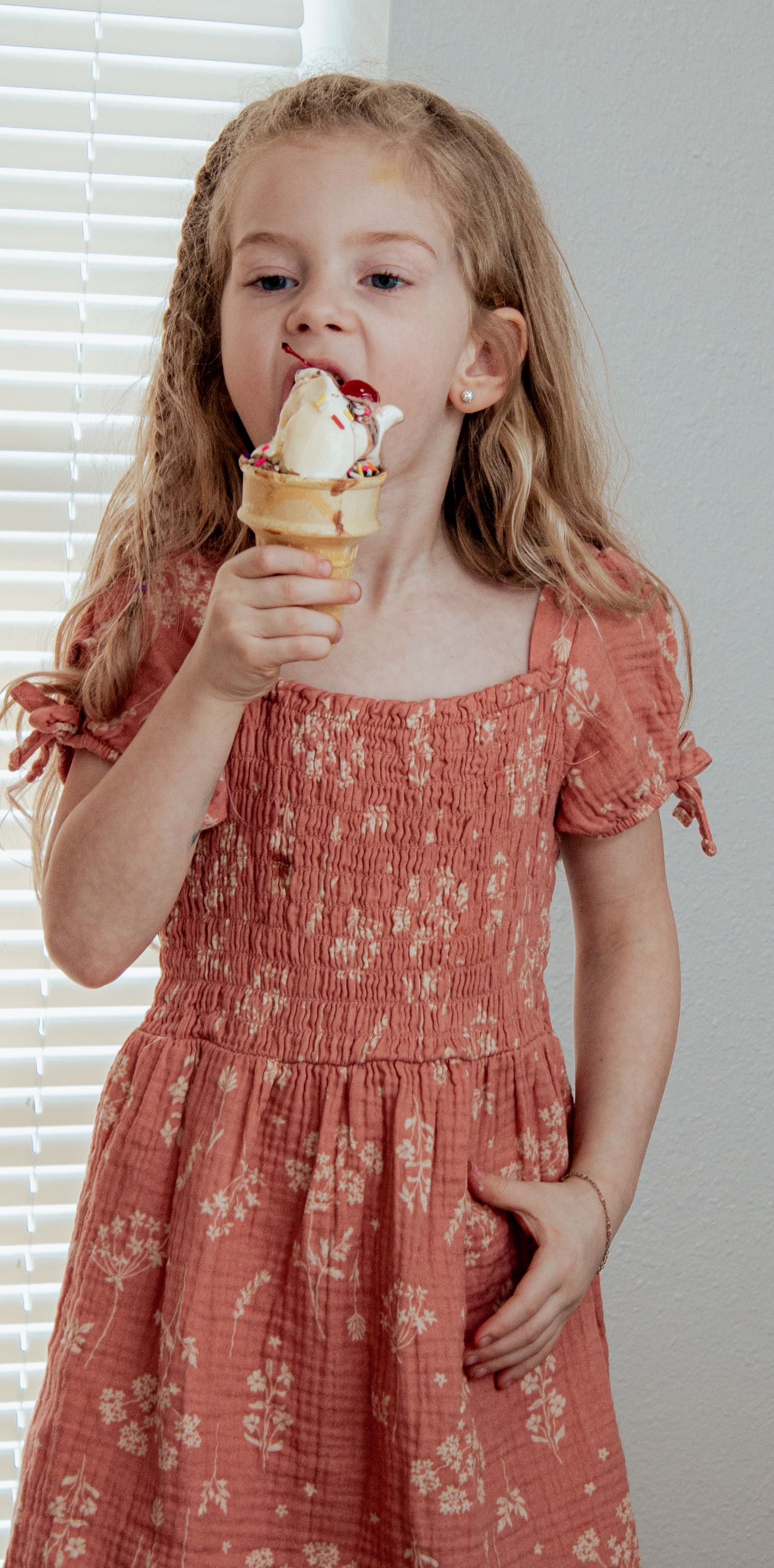 A young child in a coral floral dress enjoys eating an ice cream cone while standing against a white wall with blinds.
