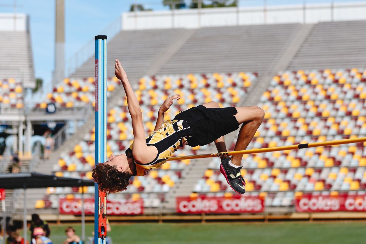 High jumper arches backwards over crossbar during competitive track and field event at stadium with colorful seating.