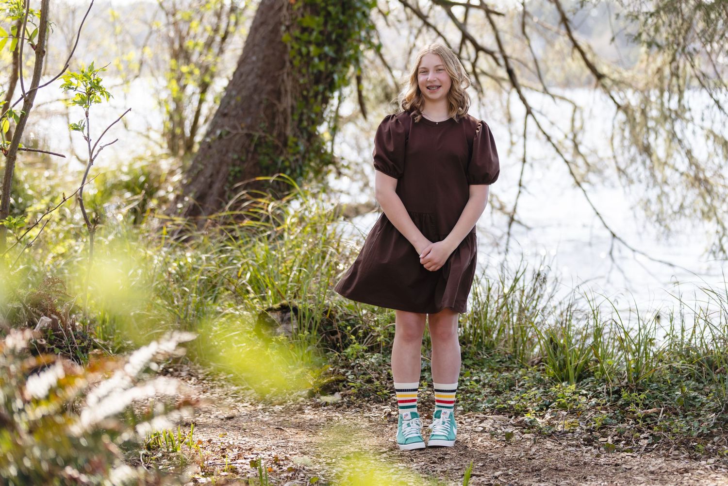 Woman in brown dress posing on forest path wearing aqua blue sneakers beside a lake on a sunny day.