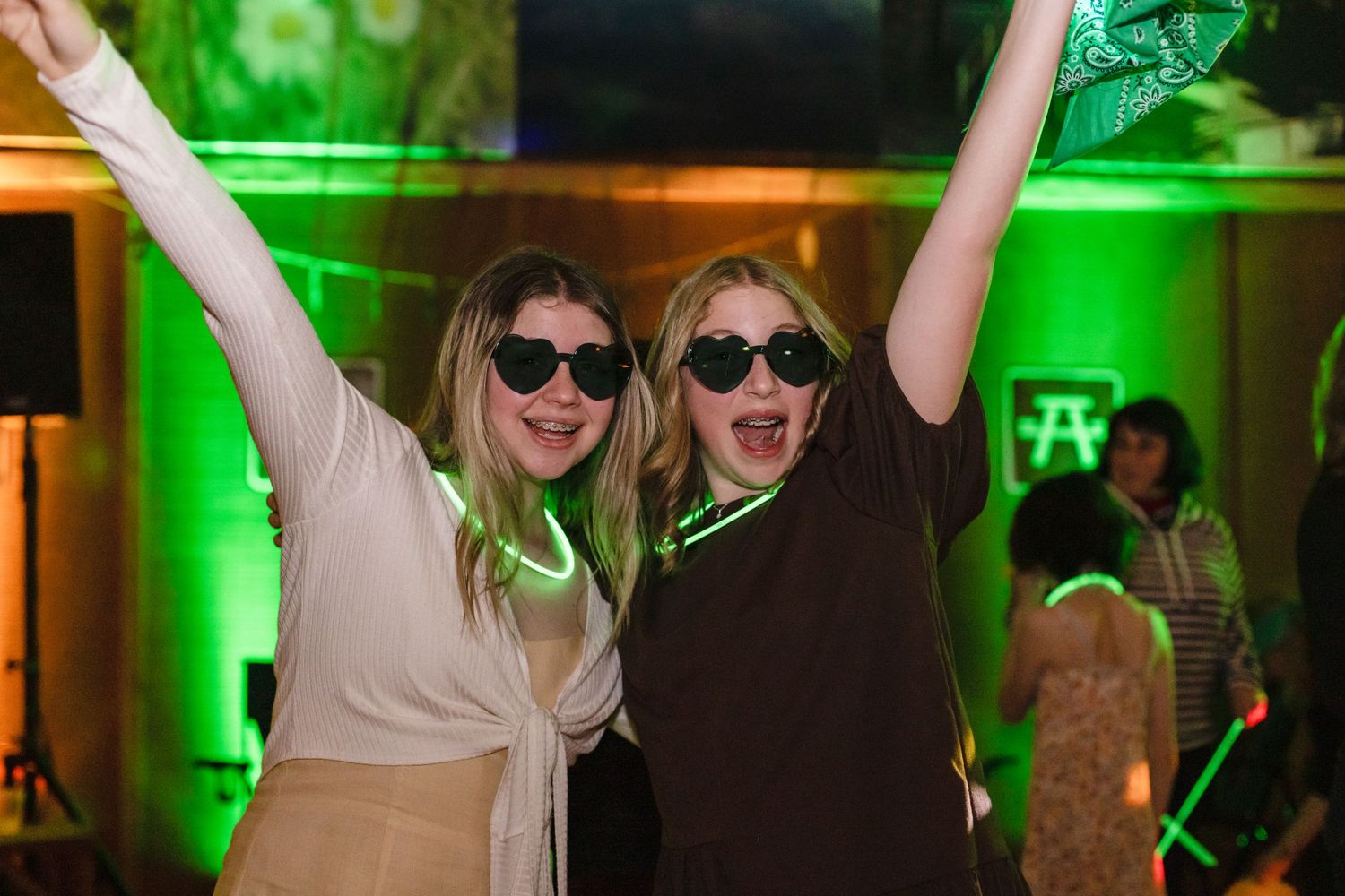 Two people in sunglasses dance with raised arms at a party with green lighting in the background.