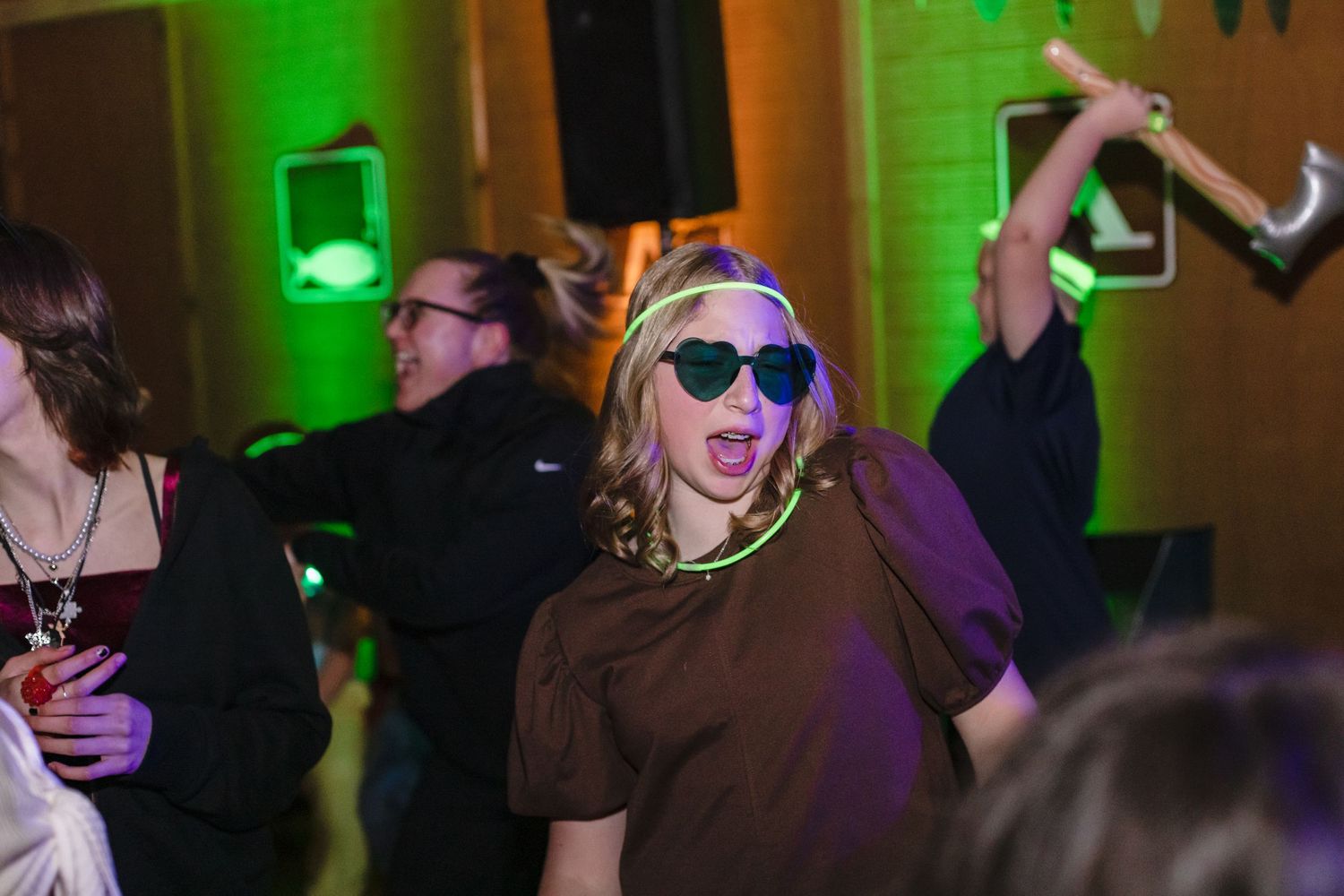Partygoer in sunglasses and brown t-shirt dancing energetically under green lighting at a nighttime event.