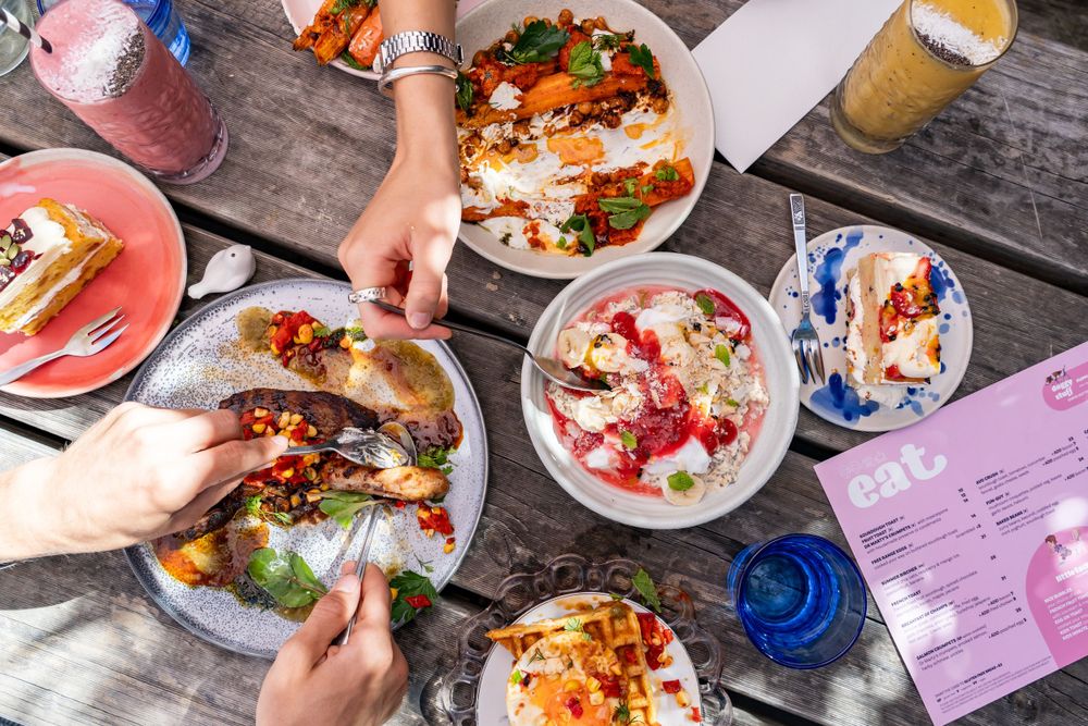 Overhead view of hands reaching for colorful plates of food and drinks on wooden table during casual dining gathering.