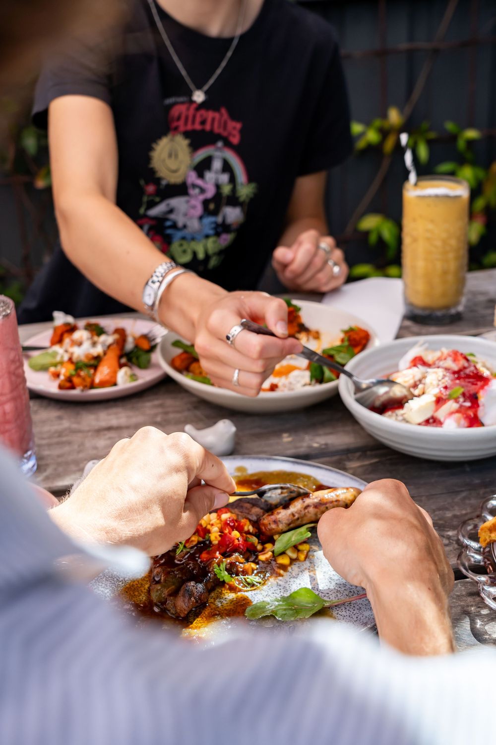 People enjoying a casual dining experience with plates of colorful food and a smoothie on a wooden table.