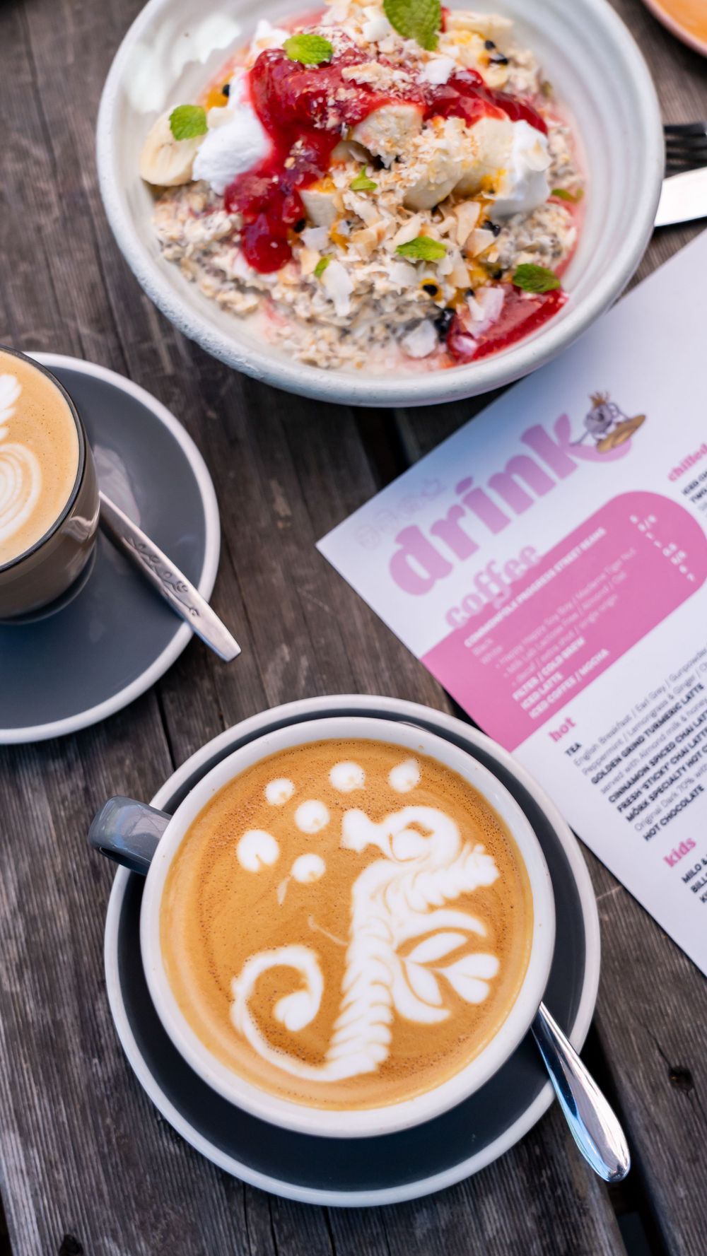 Artful latte art featuring a dinosaur design alongside a bowl of oatmeal with berries and a pink menu on a wooden table.