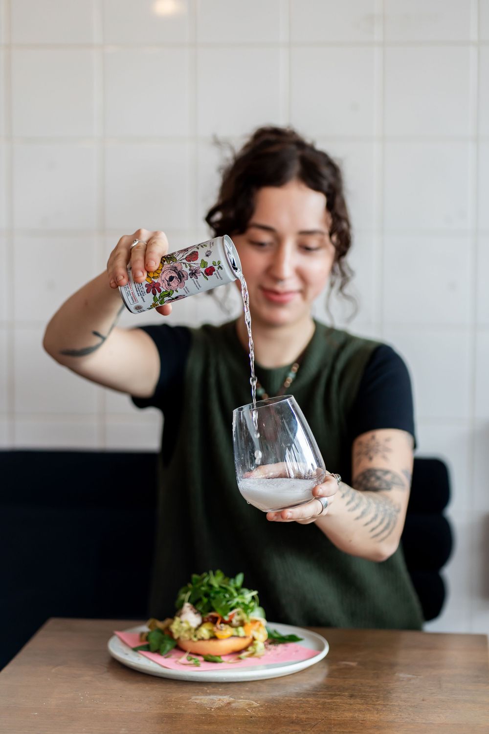 A person pours a drink from a decorative pitcher into a glass while a plated meal sits on the wooden table below.