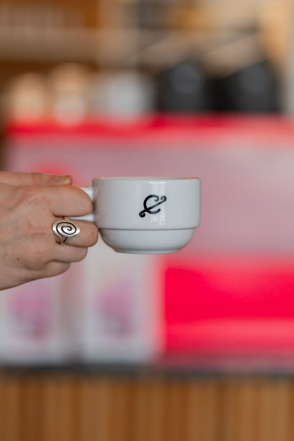 A hand holding a white ceramic coffee cup with a black logo against a blurred red background.