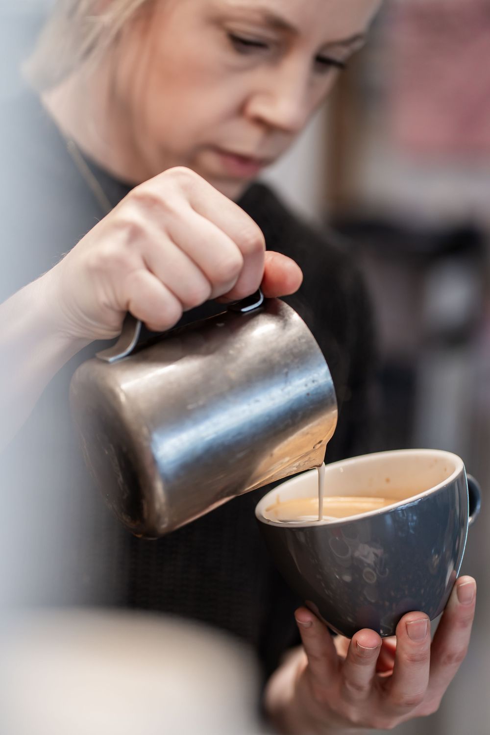 A barista pours steamed milk from a metal pitcher into a dark ceramic coffee cup creating latte art.