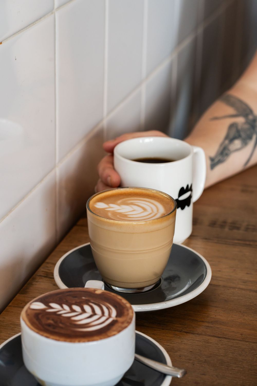 Three artfully crafted lattes with latte art on wooden table, one with black coffee mug showing visible tattoo in background.