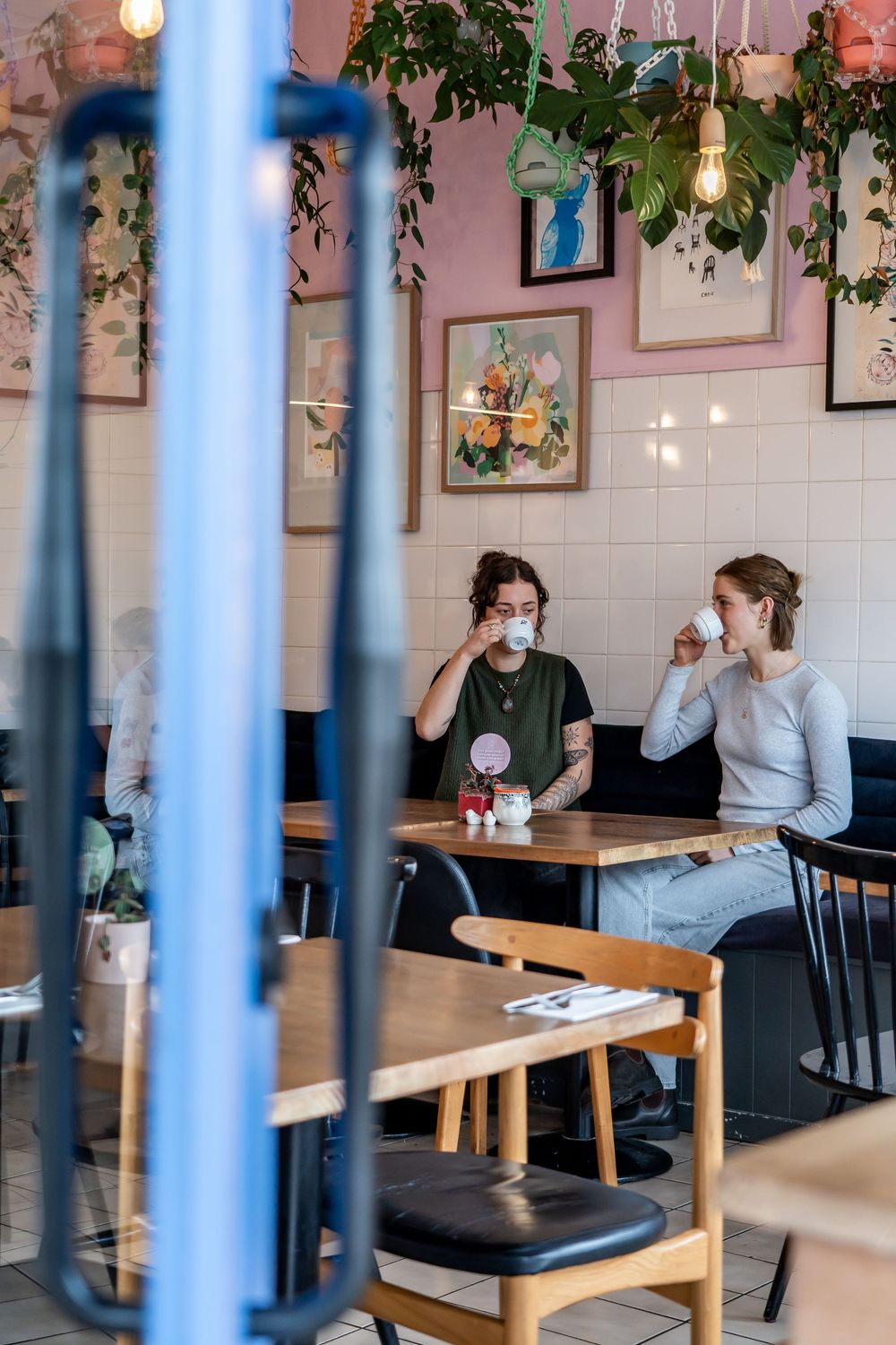Modern cafe interior features diners seated at wooden benches with pink walls, white tiles, and hanging greenery decor.