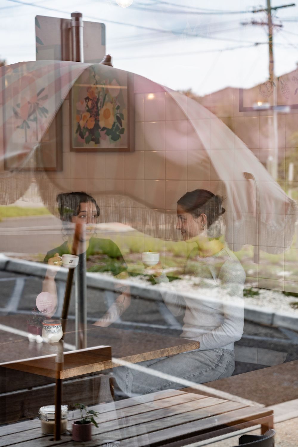 View through restaurant window showing wooden table and reflection in glass with art on wall visible in background.