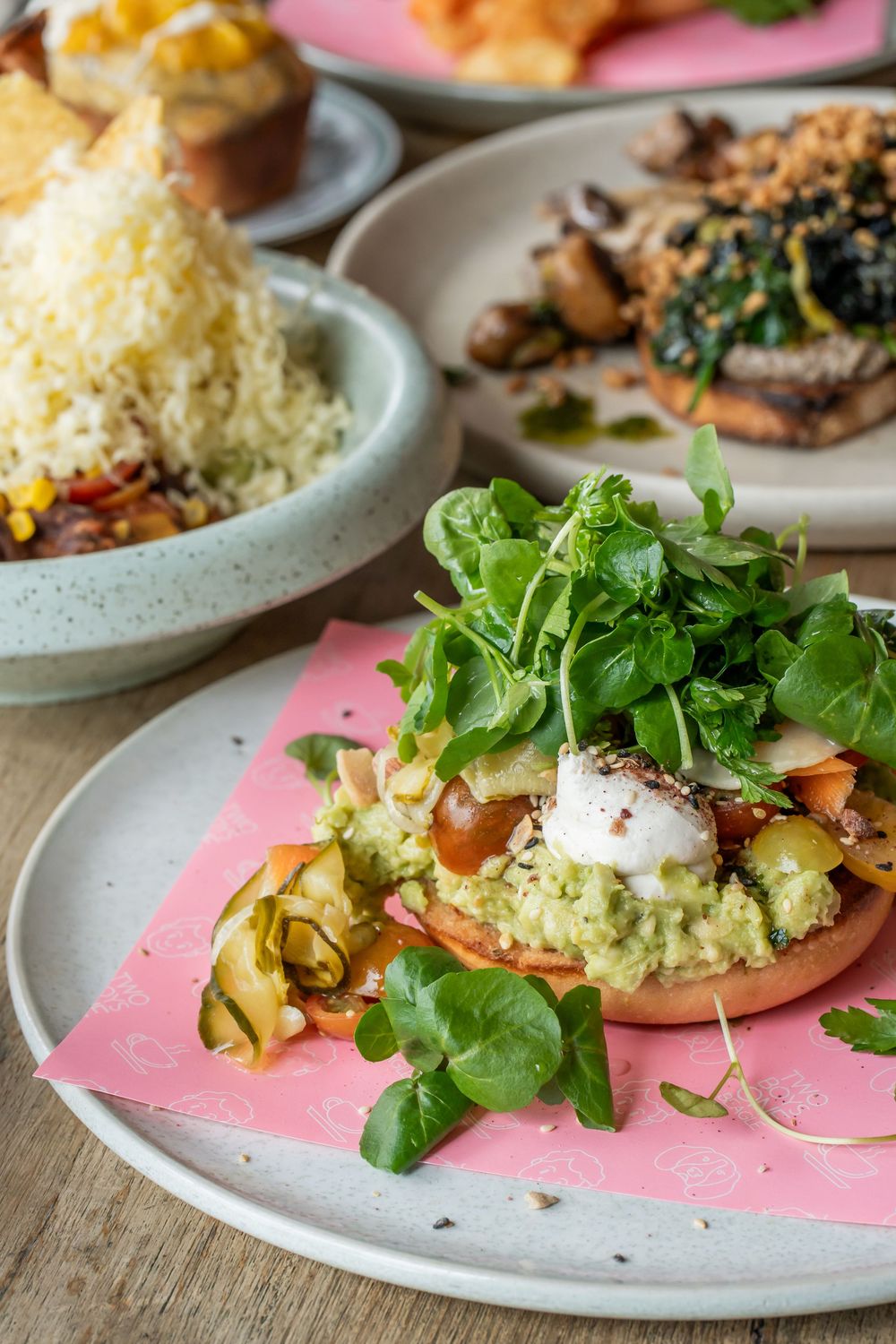 Fresh avocado bagel topped with microgreens, poached egg and served alongside mixed greens on a speckled plate.