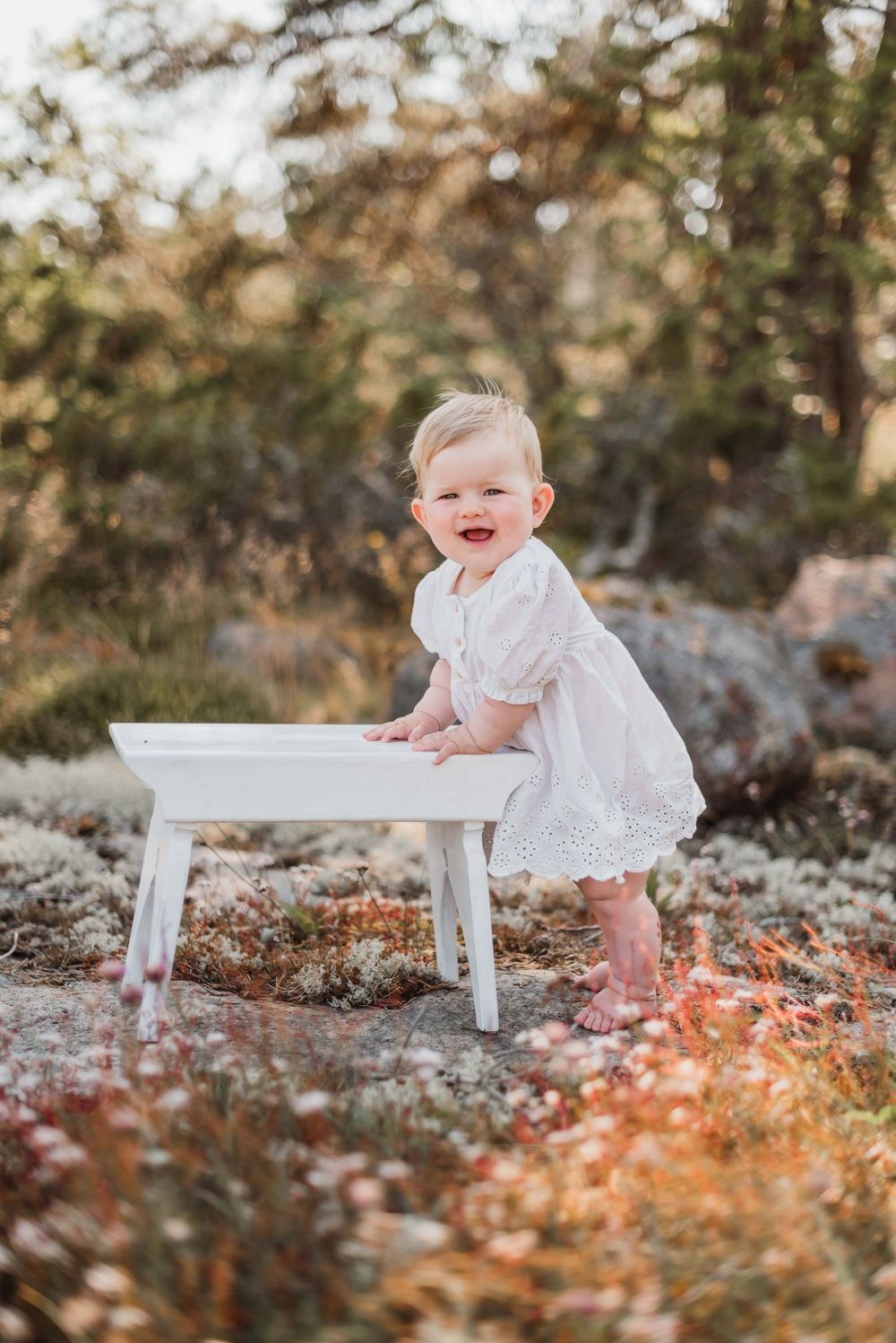 A small child in a white dress stands near a rustic white bench surrounded by autumn foliage and flowers.