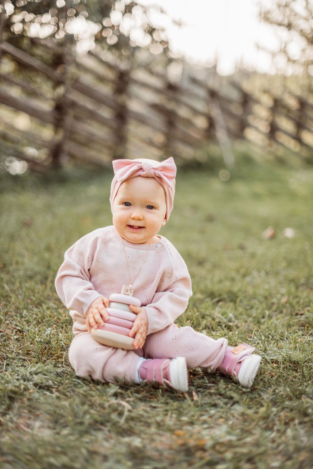 A baby in a pink outfit sits on green grass during a sunny day outdoors.