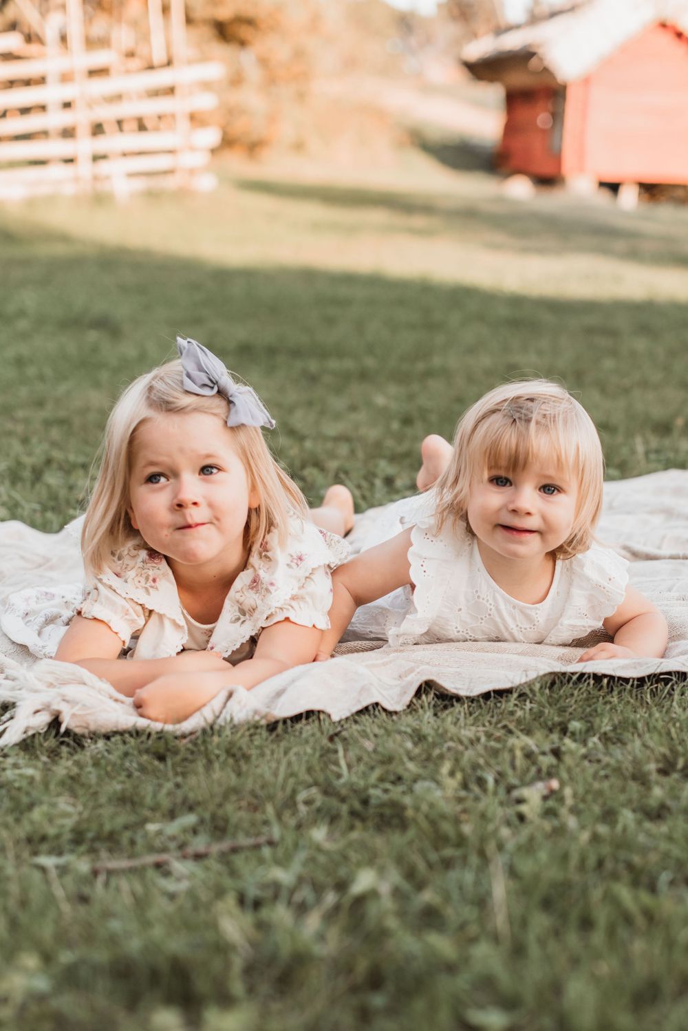 Two young children in white lace dresses lying on a blanket in grass with a red barn in the background.