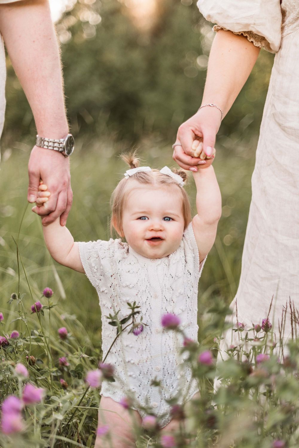 Baby in white dress holding parents' hands while standing among purple wildflowers.