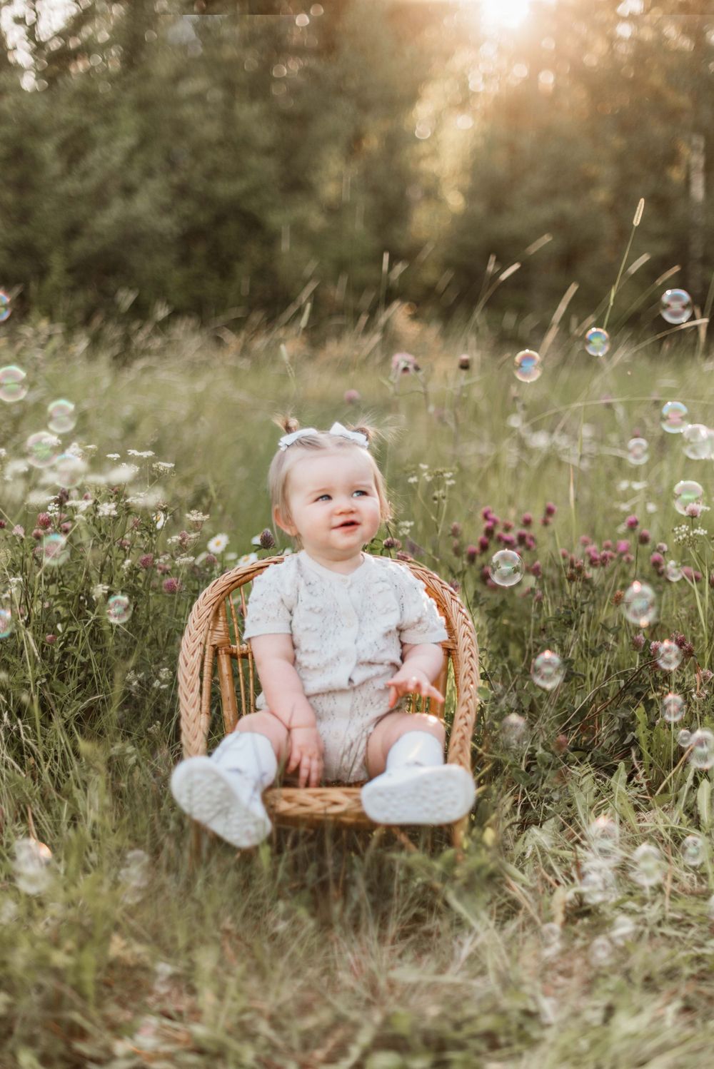 Baby sitting in wicker chair surrounded by wildflowers and bubbles at sunset.