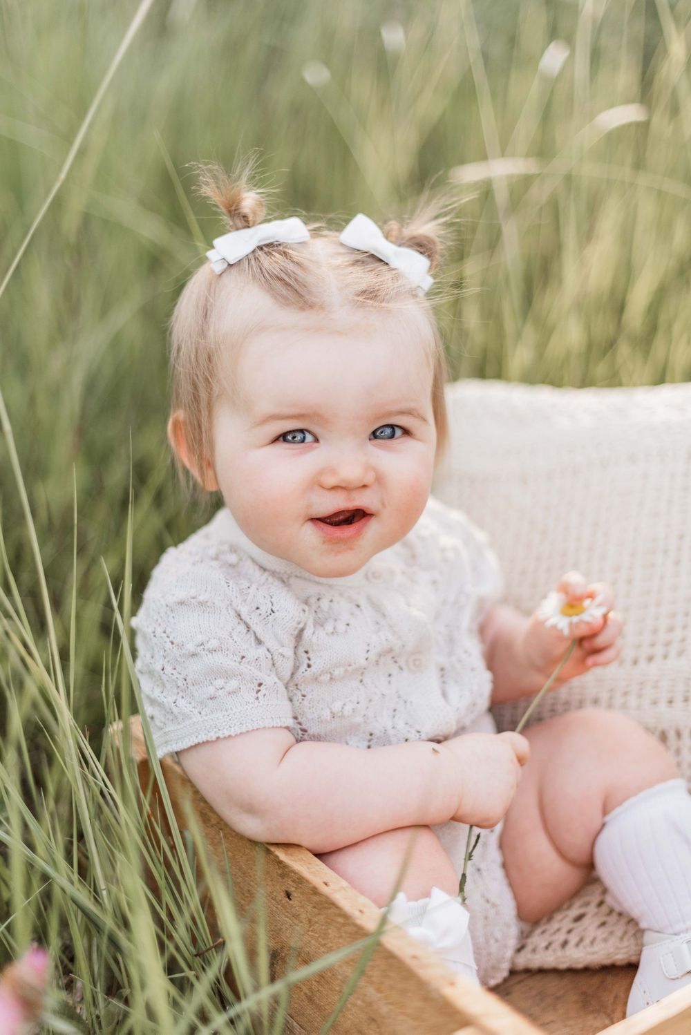 Baby in white lace dress sitting on chair in tall grass with soft natural lighting.