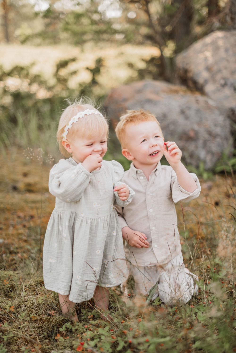 Two young children in light colored linen outfits stand together outdoors on an autumn day.