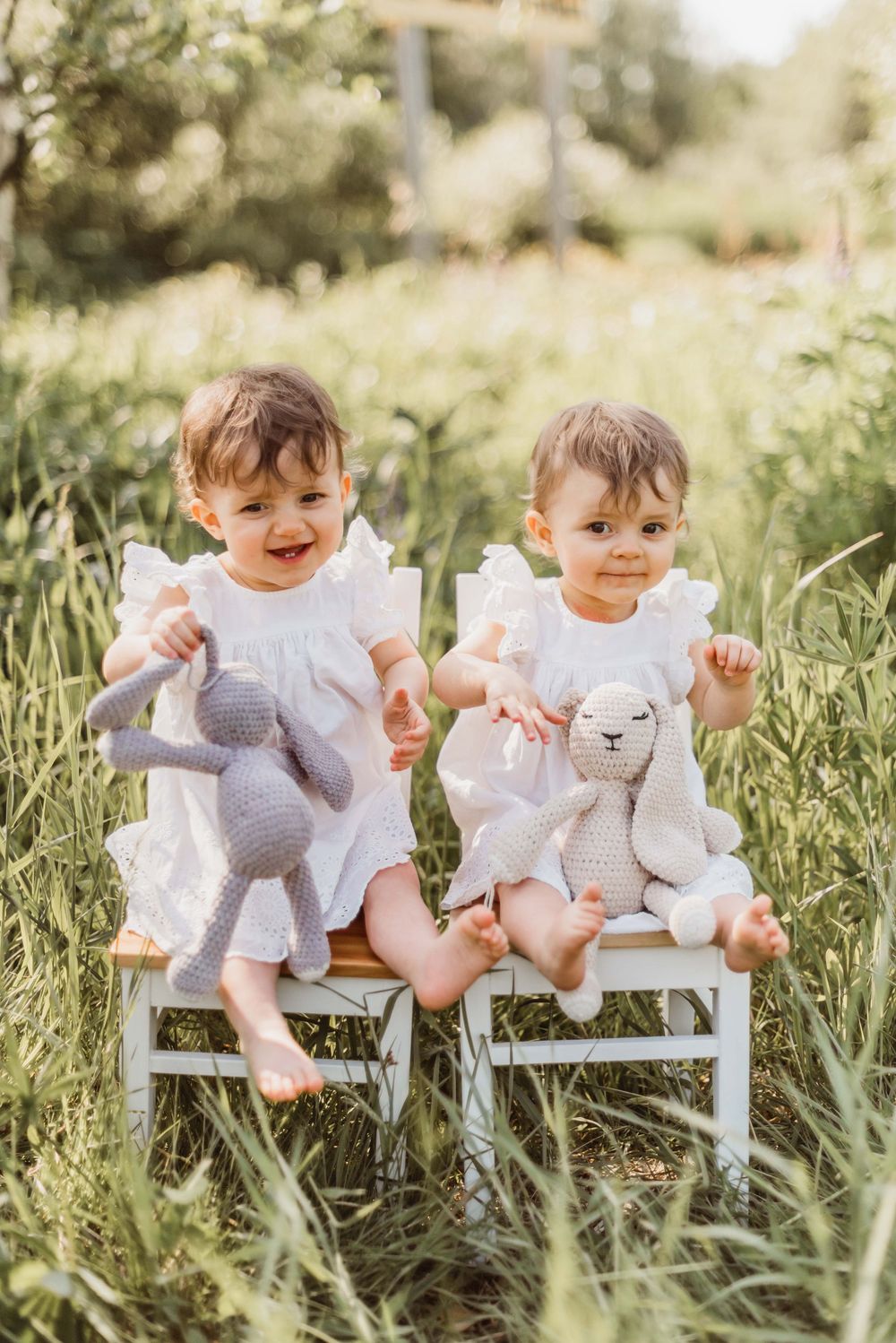 Two toddlers in white outfits sit on small chairs surrounded by tall grass in a sunny outdoor setting.