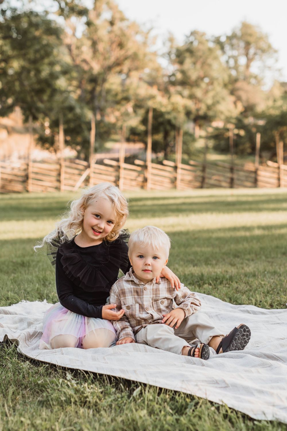Two children relax together on a white blanket in a sunny park setting during golden hour.