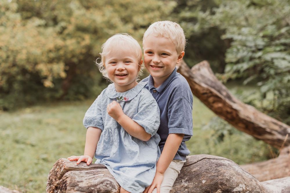 Two children sit together on a fallen tree trunk in a lush green outdoor setting.