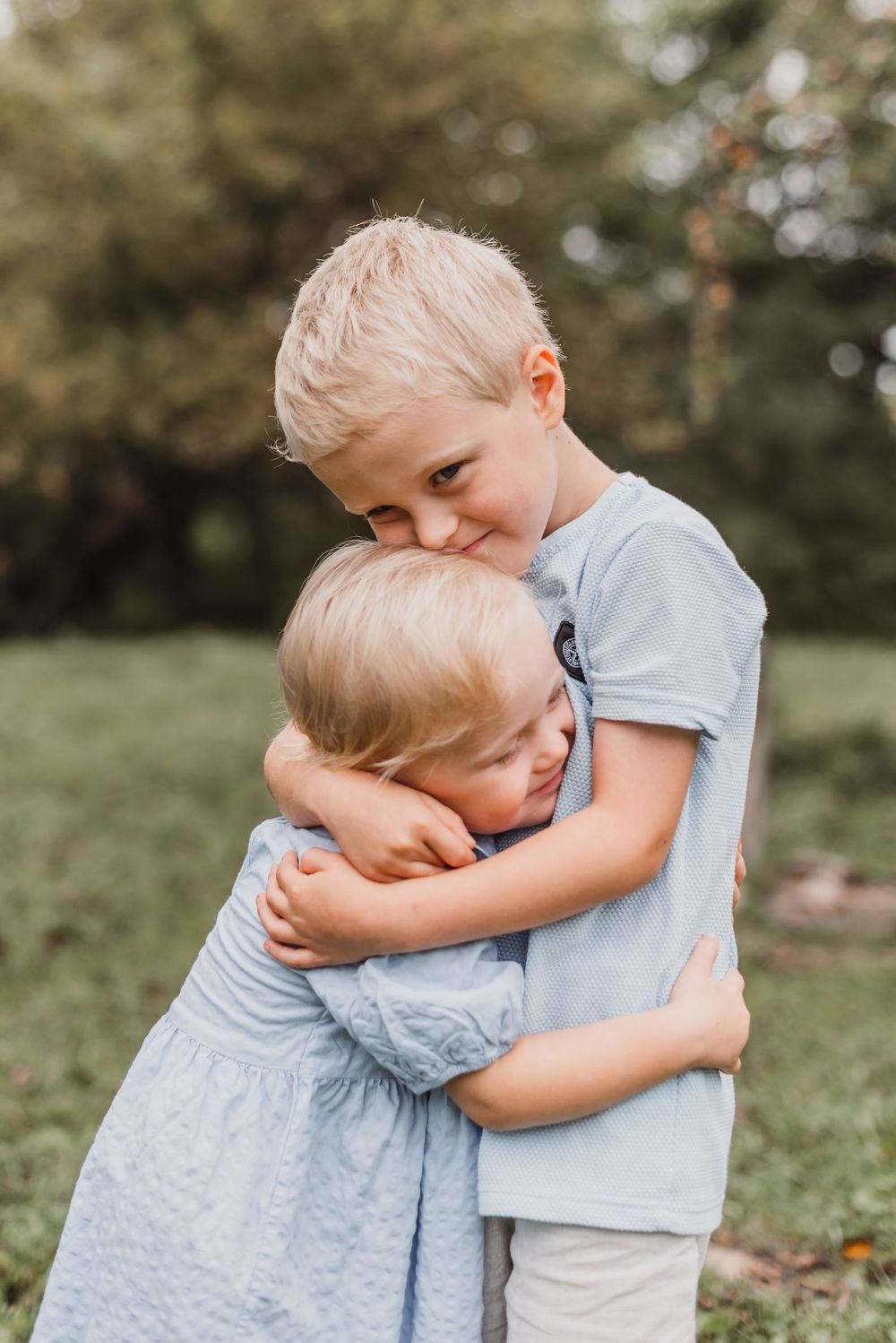 Two children share a tender embrace outdoors in a grassy field with soft bokeh lighting in the background.