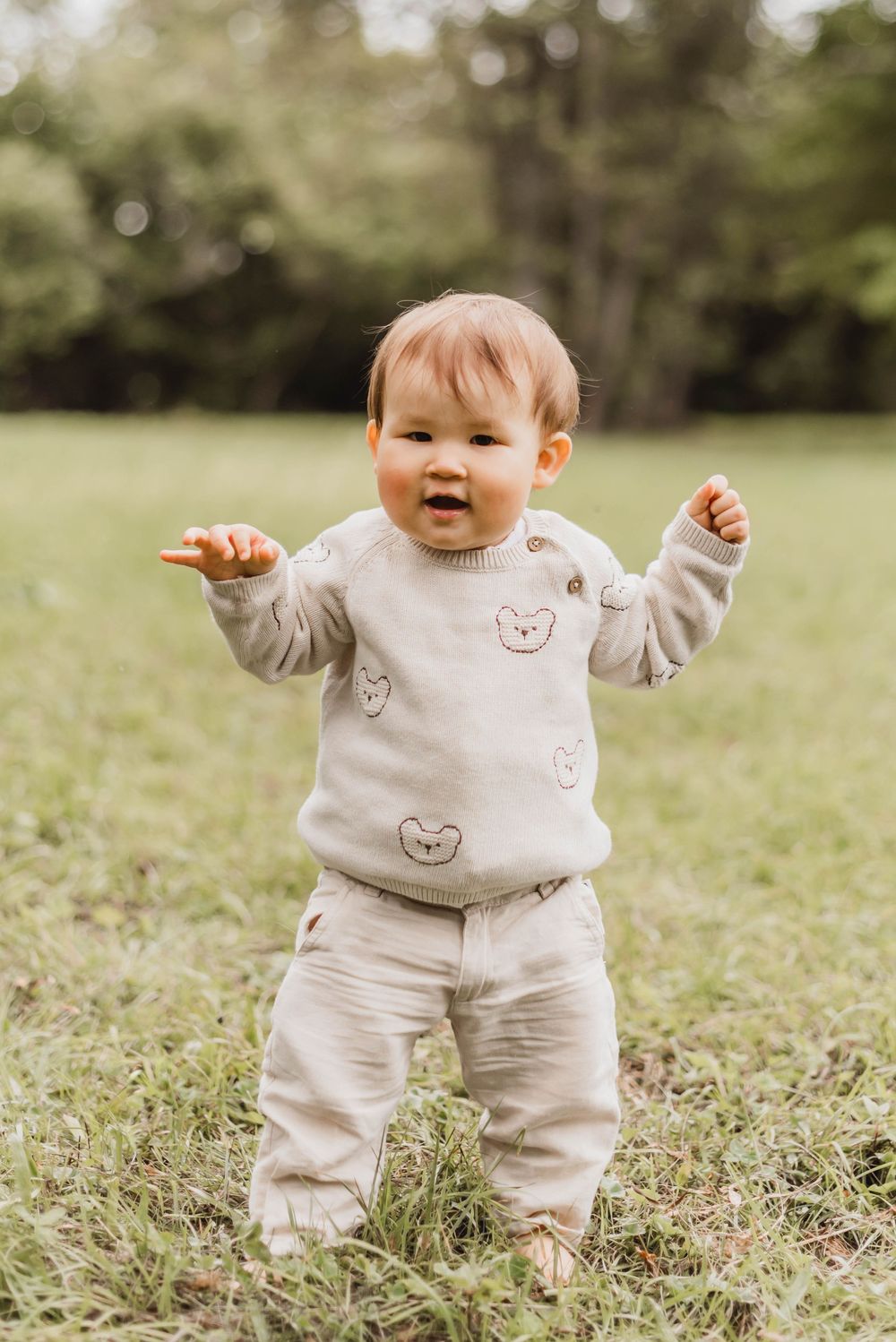 A cheerful baby wearing a beige sweater with bear prints and matching pants stands in a grassy field on a sunny day.