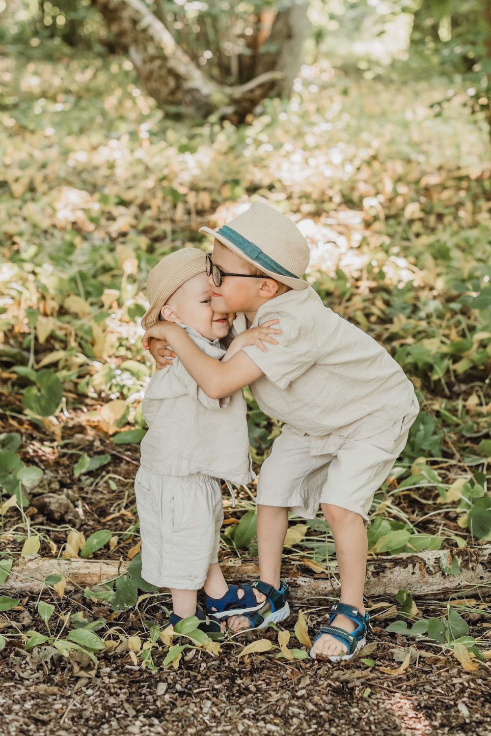 Two young children in light colored clothing share a sweet moment while playing together outdoors in a garden setting.