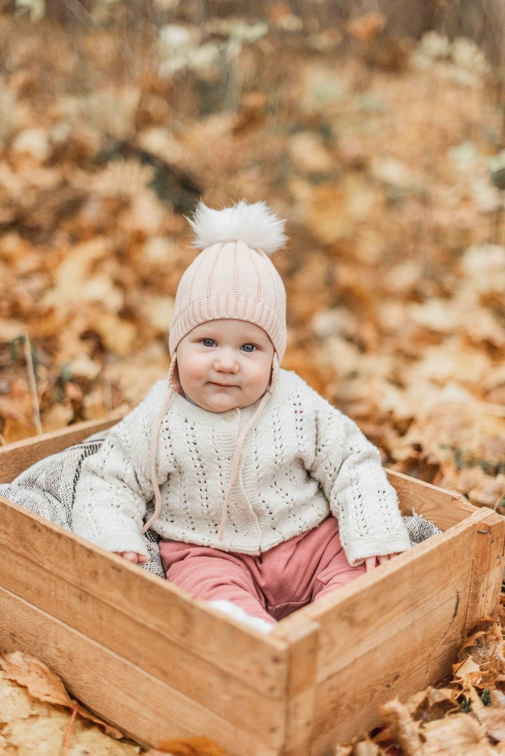 A baby in a white knit sweater and pink hat sits in a wooden crate surrounded by autumn leaves.