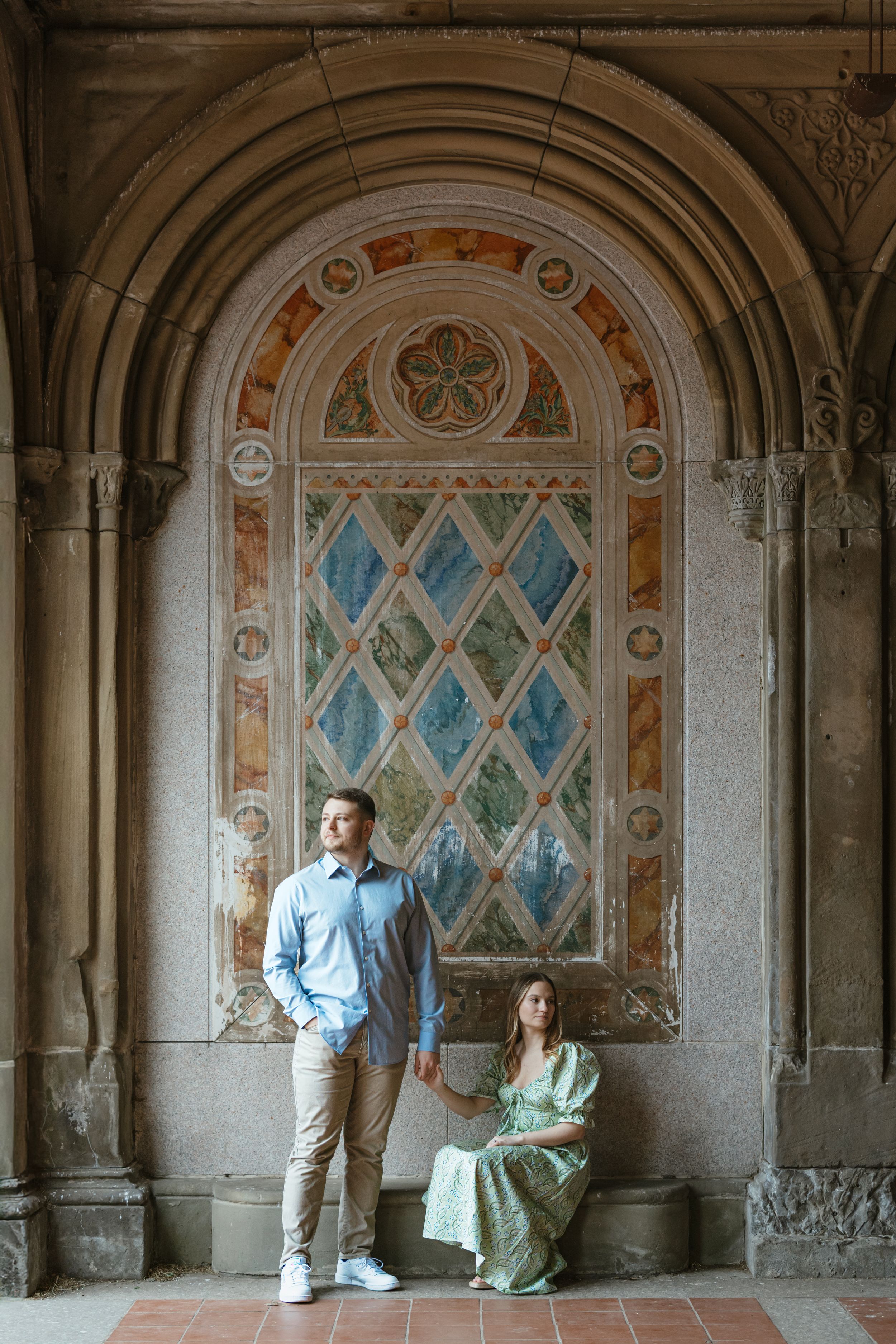 Two people pose under an ornate stone archway with blue diamond-patterned stained glass and decorative architectural details.