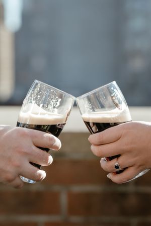 Two hands holding dark beer glasses coming together in a celebratory toast with foam and bubbles visible at the top - Engagement Photography