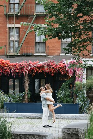 A couple embraces and shares a romantic moment on a stone wall outside a brick building with blooming flowers.