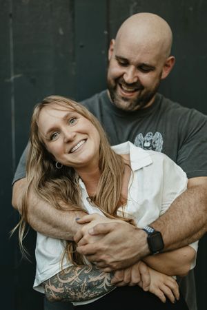 A loving couple embraces and smiles in front of a dark wall, with natural lighting and casual attire -Engagment Photo - Hudson Valley