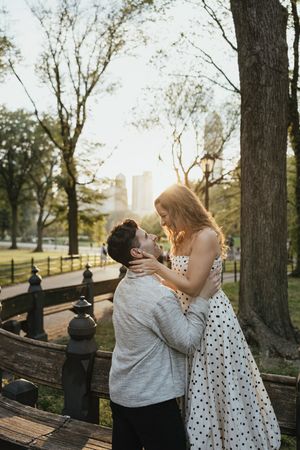 A romantic couple shares an intimate moment on a park bench at sunset with city buildings visible in the background.