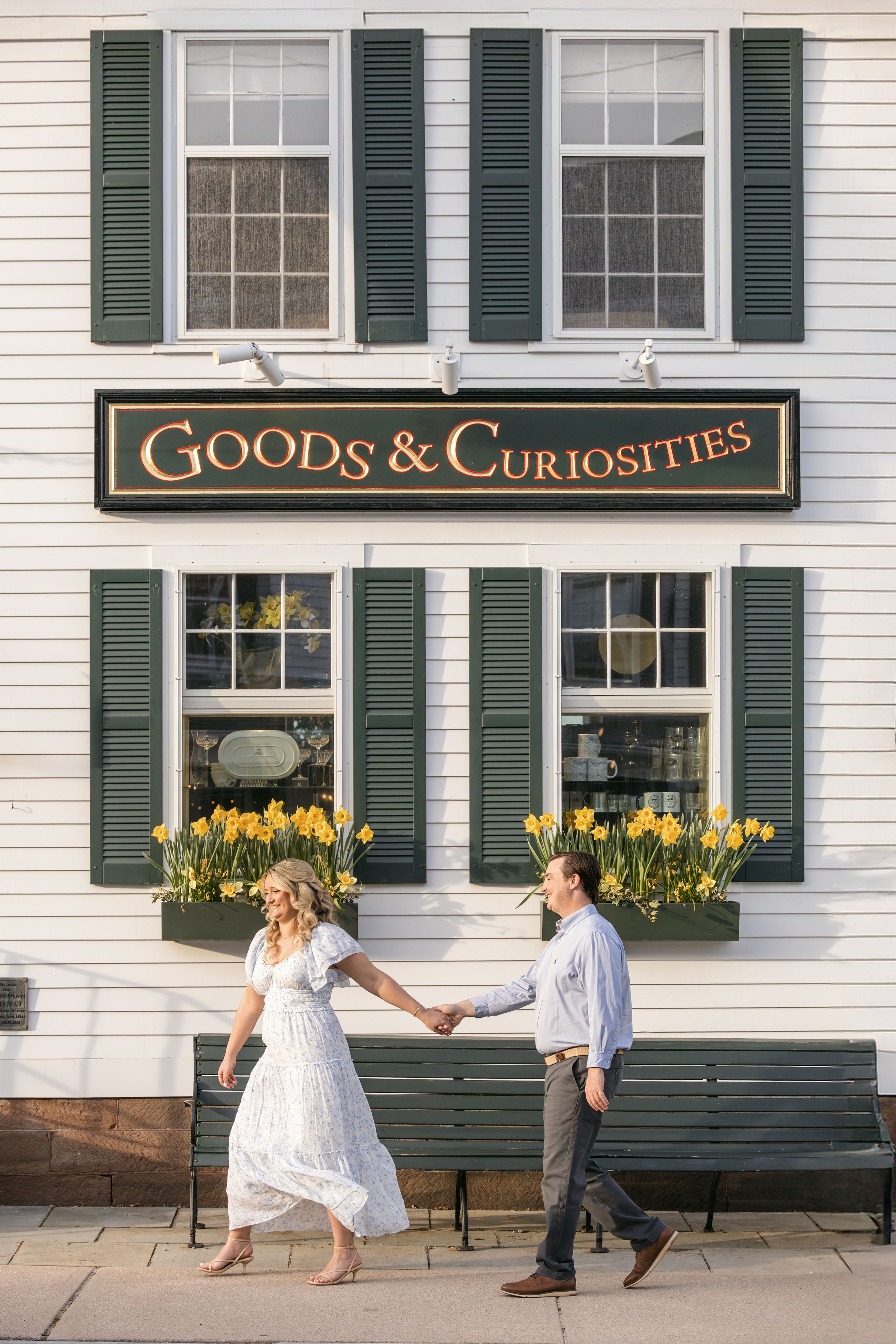A couple holds hands in front of a white colonial building with green shutters and a 'Goods & Curiosities' storefront sign.