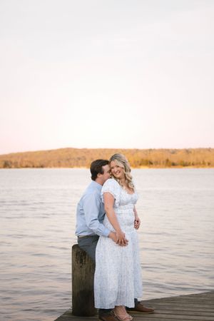 Romantic couple in white and blue outfits share a tender moment on wooden dock by lake at sunset.