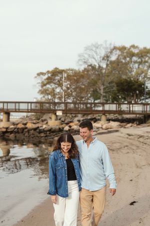 A couple walks along a rocky beach with a wooden pier and autumn trees in the background on an overcast day.