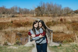 Two people embrace while standing in a grassy field with bare trees and blue sky in the background during autumn. -Engagment Photo - Hudson