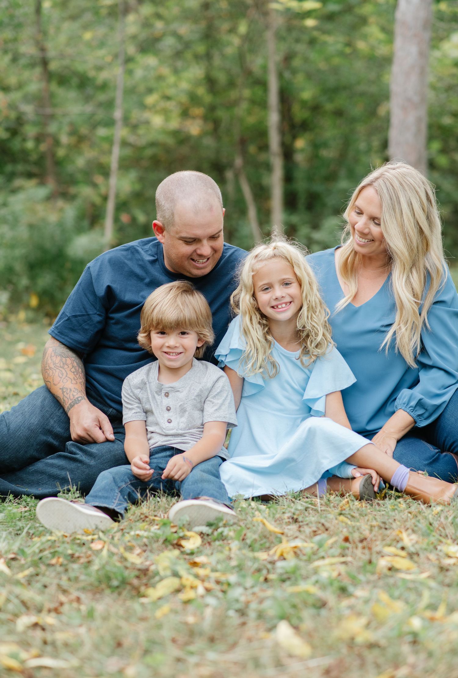 Family sits together on fallen autumn leaves wearing coordinating blue and grey outfits in an outdoor woodland setting.