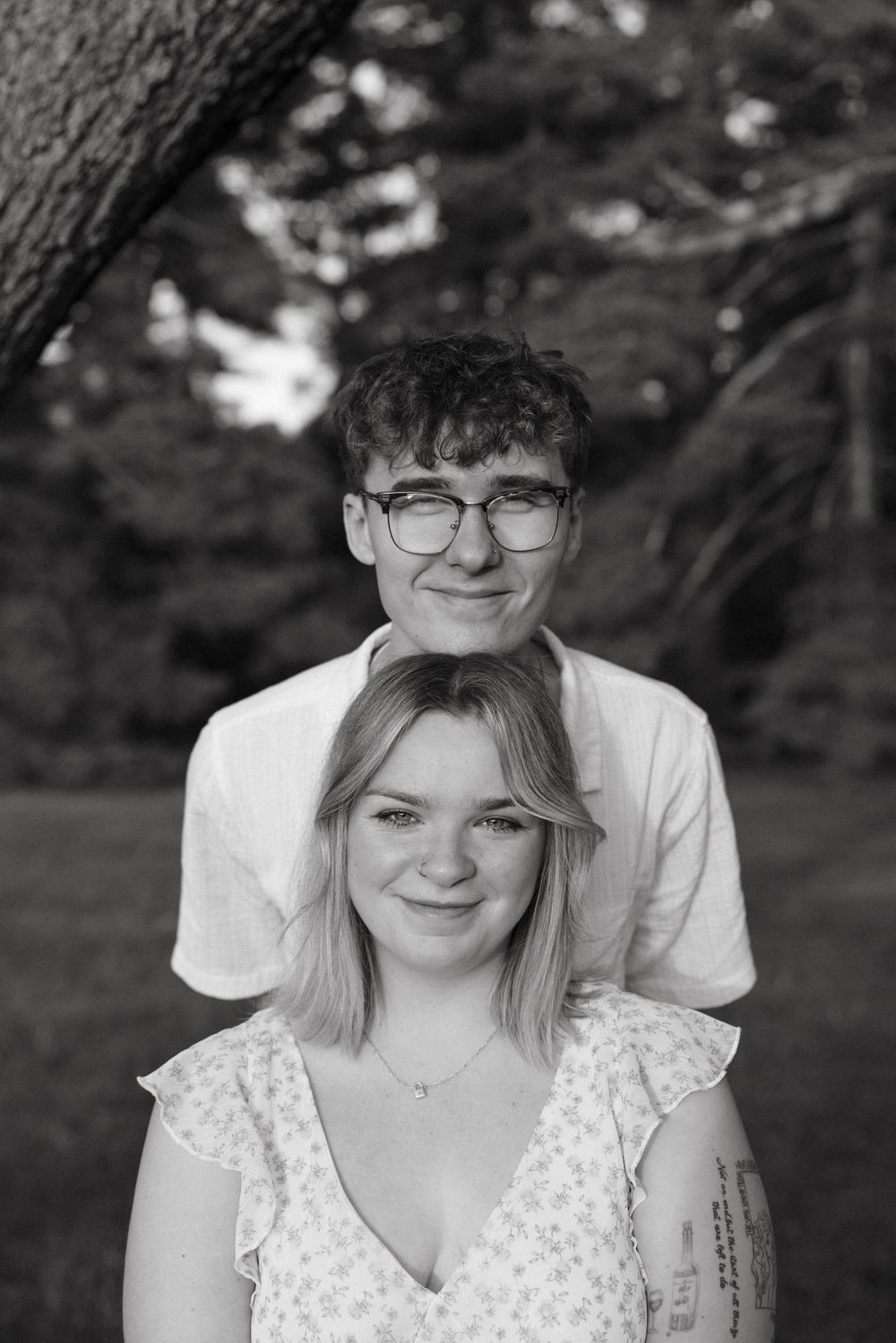 Black and white portrait of a young couple smiling outdoors in a park with lush trees in the background.