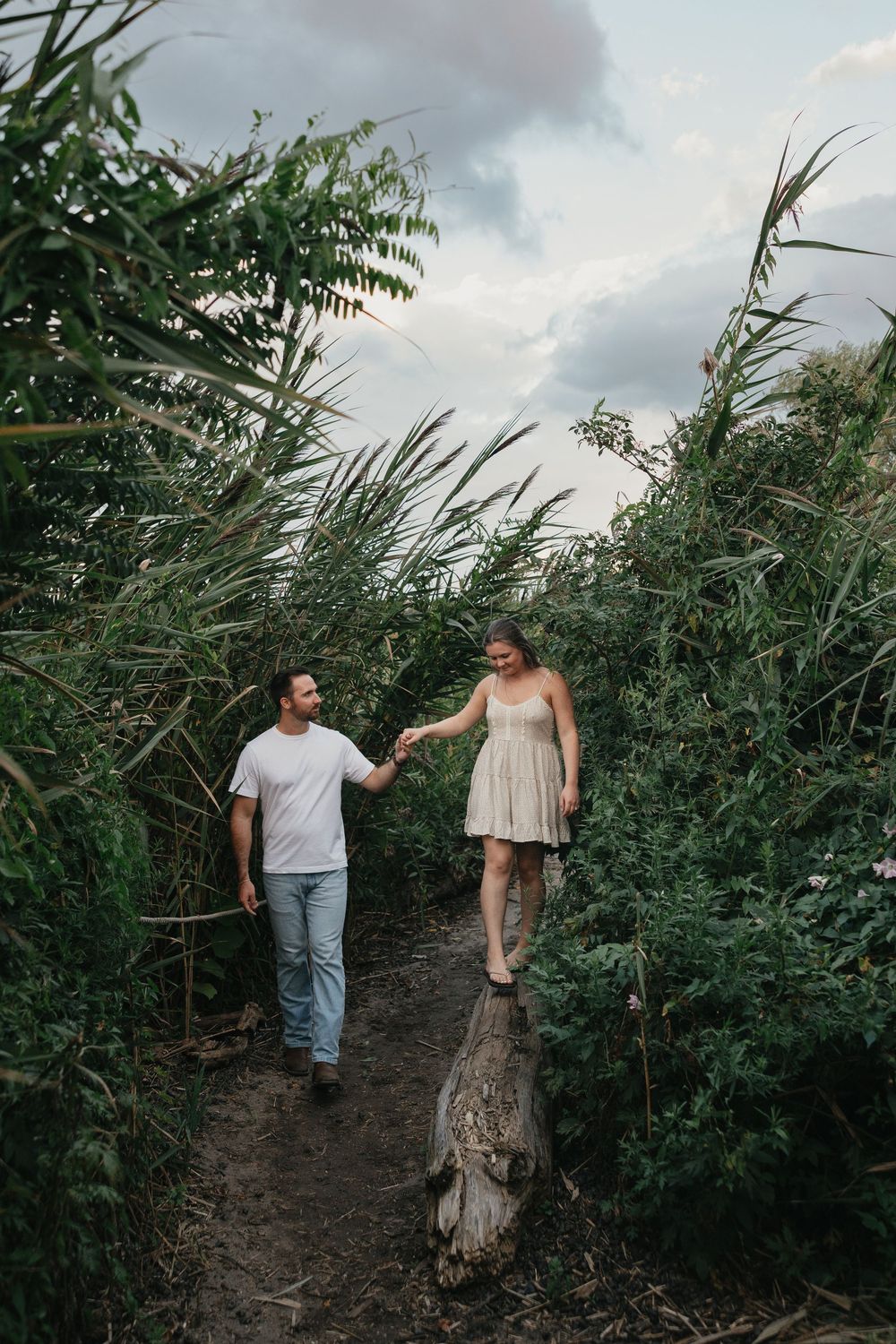 Couple holding hands walking through lush green reeds on a dirt path, woman in white dress balancing on a log.