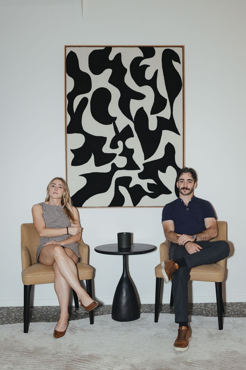 Two people seated in tan chairs before a bold black and white abstract painting in a modern interior.