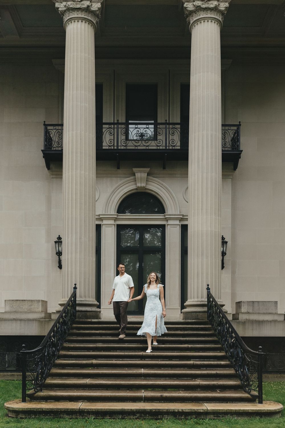Couple holding hands walking down grand staircase of elegant neoclassical mansion with tall columns.