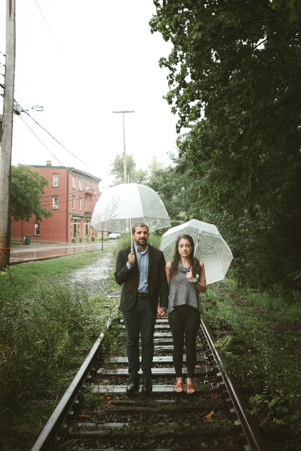 Couple holding clear umbrellas standing on rain-soaked railway tracks surrounded by lush green trees and a red brick building.
